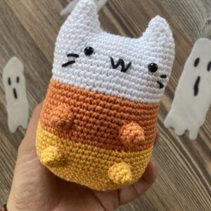 Una mano sostiene el Candy Corn Cat - Gato Halloween Amigurumi, un juguete de peluche tejido a crochet con colores de caramelo de maíz, mientras que en el fondo se ven recortes de fantasmas.