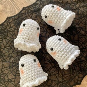 Cuatro figuras amigurumi de crochet "Fantasma", con ojos negros y mejillas sonrosadas, están dispuestas sobre una tela de encaje negro con un diseño de telaraña.